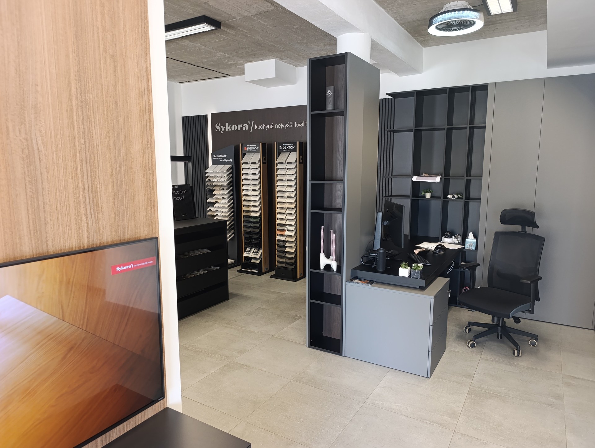 Interiér kuchyňského studia s vzorky materiálů, pracovním stolem a černou židlí v šedém provedení